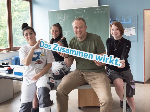 Schülerinnen mit Lehrer halten gemeinsam ein "Das Zusammen wirkt" Schild.