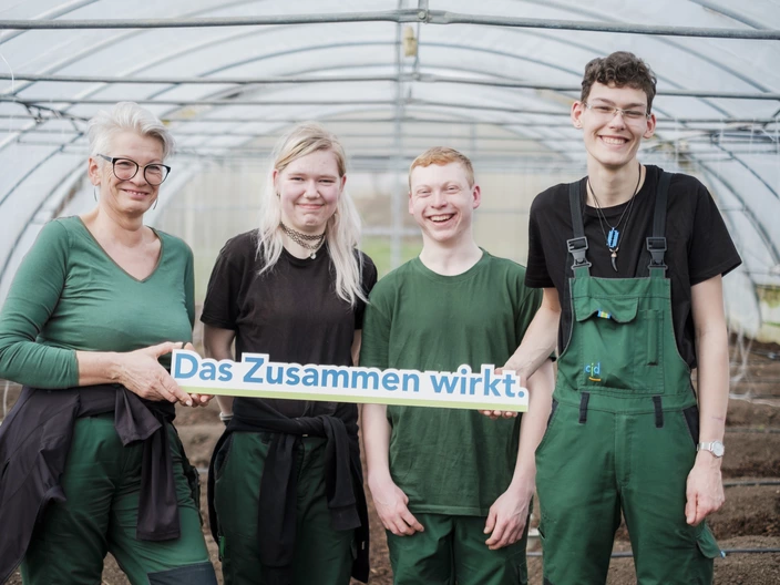 Drei Gartenbau-Auszubildende halten ein "Das Zusammen wirkt"-Schild mit ihrer Ausbilderin 