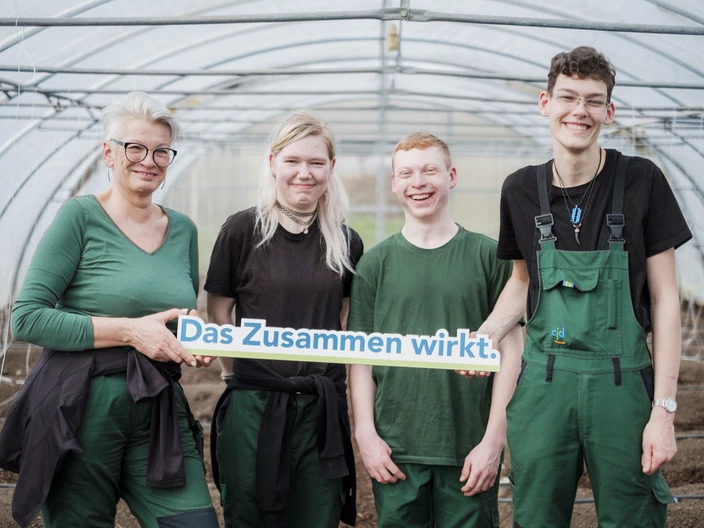 Drei Gartenbau-Auszubildende halten ein "Das Zusammen wirkt"-Schild mit ihrer Ausbilderin 