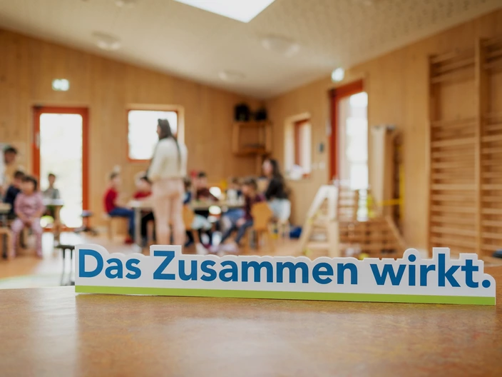 "Das Zusammen wirkt"-Schild mit einer Gruppe an Menschen im Hintergrund