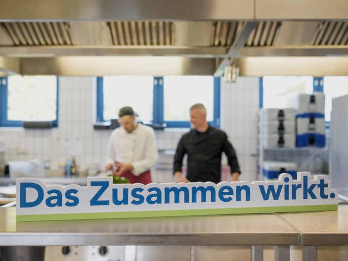 "Das Zusammen wirkt"-Schild mit zwei Köchen im Hintergrund